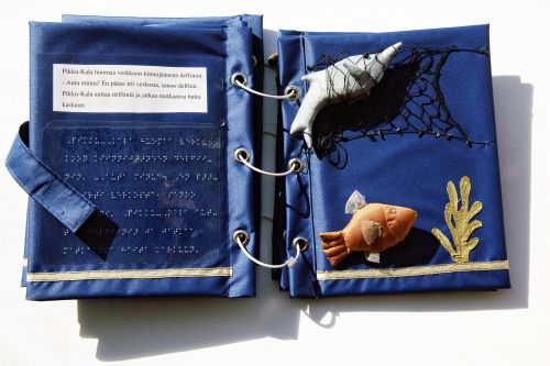 En uppslagen bok överdragen med blått tyg. På den högra sidan finns en tredimensionell orange fisk och ovanför den en grå delfin i ett nät. På den vänstra sidan finns text i svartskrift och punktskrift