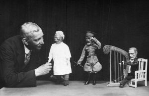 Mustavalkokuva miehestä kolmen nuken kanssa.