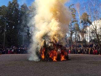 Villisti savuava ja palava kokko, ihmisiä kaukaisuudessa katsomassa.