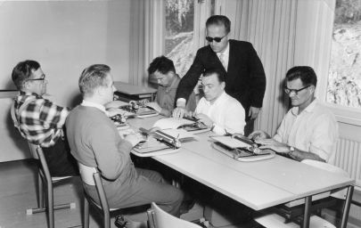 Black-and white photo where people are sitting by a desk. One person is standing up teaching them.