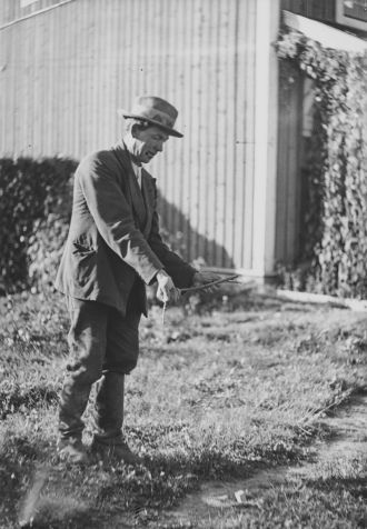 Vilttihattuinen mies kävelee nurmikolla ja pitää käsissään haarakasta puunoksaa. Taustalla on pystyvuorattu rakennus. Kuva Museovirasto, kuvaaja Pekka Kyytinen, 1930–1939.