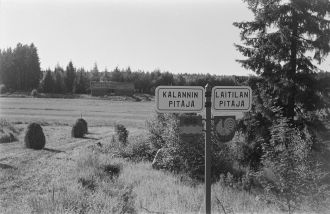 Kyltit ja vaakunat maalaismaisemassa, taustalla heinäseipäitä.