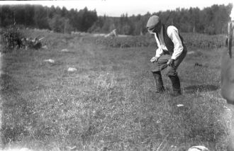 Mustavalkokuvassa käsivartensa polviin tukenut mies pitää sormissaan haarakasta pajunoksaa pellolla. Kuva: Pielisen museo.