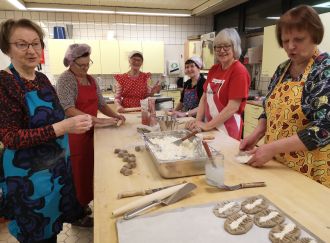 Joukko ihmisiä seisoo talouskeittiössä pöydän ääressä leipomassa karjalanpiirakoita.