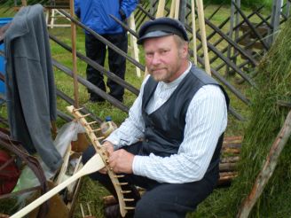 Hattuun, liiviin ja kauluspaitaan pukeutunut mies istuu rakentamassa puista haravaa.