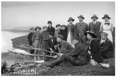 Mustavalkoinen valokuva, jossa joukko ihmisiä on joen rannalla. Muutama heistä istuu etualalla paistamassa varraslohta. Kuvan vasemmassa alalaidassa lukee teksti Rovaniemi lohenpaisto.