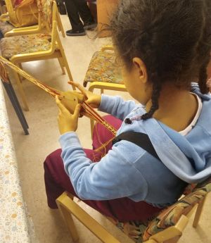 Kuvassa lapsi istuu tuolilla ja kutoo lautanauhaa historianelävöitysyhdistyksen käsityötapaamisessa. Kuva: Aino Hujanen