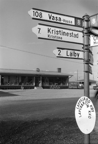 Ruotsinkielinen tiekyltti, taustalla vanha kauppa. Mustavalkokuva.