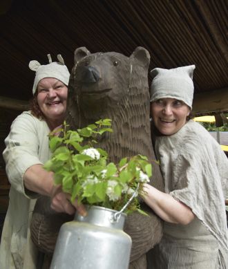 Two people in light linen capes and hats hugging a wooden bear.