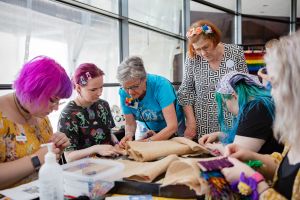 Pöydän äärelle on kumartuneina viisi eri ikäistä naista yhteistä ryijyä tekemään.