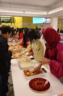 Valokuva Pitha Uthsob festivaaleilta. Pitkän pöydän ääressä seisoo joukko ihmisiä jakamassa erilaisia bangladeshilaisia leivonnaisia.