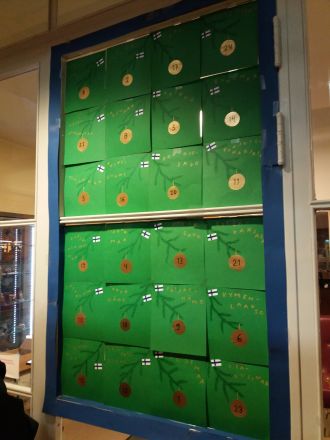 Vihreä kalenteri, joka on koristeltu Suomen lipuilla ja piirrustuksilla joulukuusenkoristeista.
