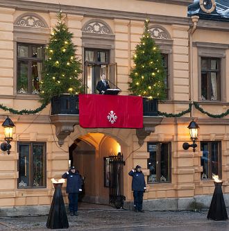 Henkilö julistaa joulurauhaa parvekkeella, jota koristaa kaksi joulukuusta.