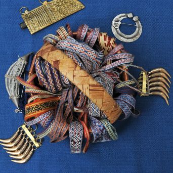 Kuvassa on ennallistuksia Suomen rautakauden lautanauhoista. Värikkäät nauhat on aseteltu koriin ja korin ympärillä on pronssisia karhunhammasriipuksia sekä puukontuppi. Rautakaudella lautanauhat saattoivat kannatella erilaisia riipuksia. Kuva: Mervi Pasanen